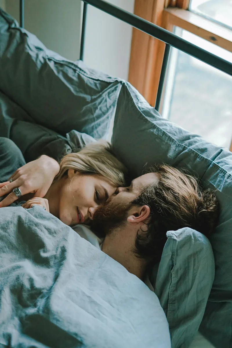 A couple lying in bed together, one partner gently kissing the other's forehead in a tender, trusting moment