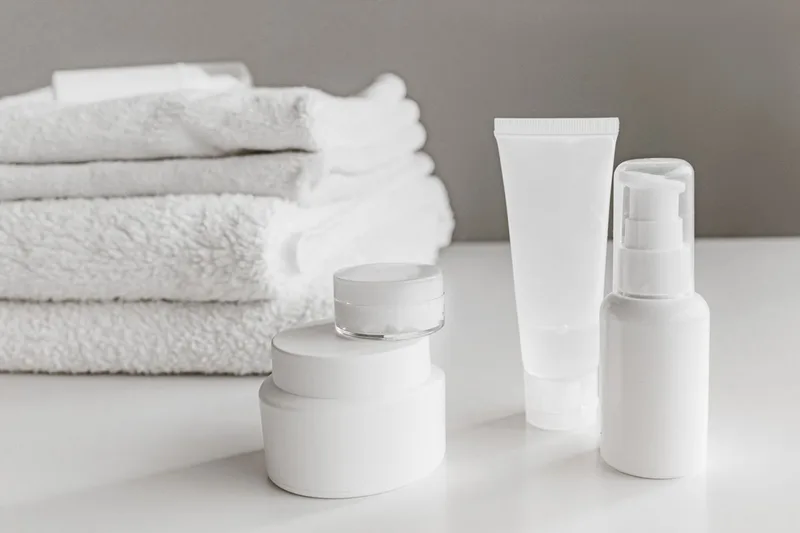 Minimalist white personal care bottles and folded towels arranged on a pale neutral surface, suggesting a simple cleaning and care routine