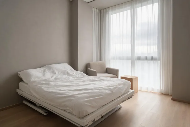 A quiet bedroom corner with a height-adjustable bed raised slightly at the head, a plain linen cover and an armless upholstered chair beside a tall window