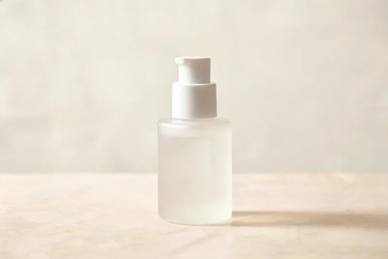 A single plain unbranded frosted pump bottle standing on a pale stone surface in soft diffused daylight