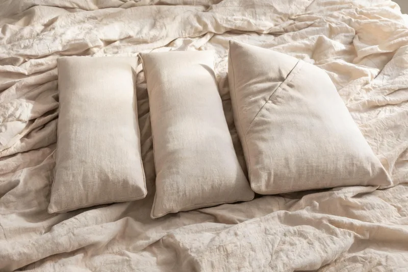 Two firm rectangular bolster cushions and a folded wedge pillow arranged on rumpled neutral linen bedding in soft directional window light