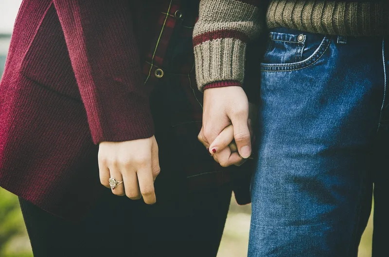 Two hands clasped together against a soft sunset meadow, a quiet non-explicit image of a couple holding hands