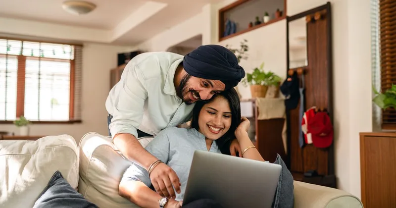 A smiling couple sitting close together on a couch at home, browsing a laptop side by side in warm indoor light, a modest non-explicit image of two people shopping together online