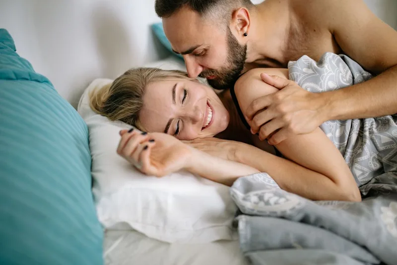 A couple laughing and cuddling under a blanket in soft morning light, a modest non-explicit image of two people relaxed together in bed