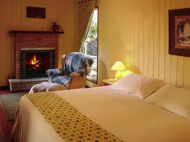 A cozy bedroom lit warmly by a fireplace and a bedside lamp, with no people present, suggesting a calm and private evening atmosphere