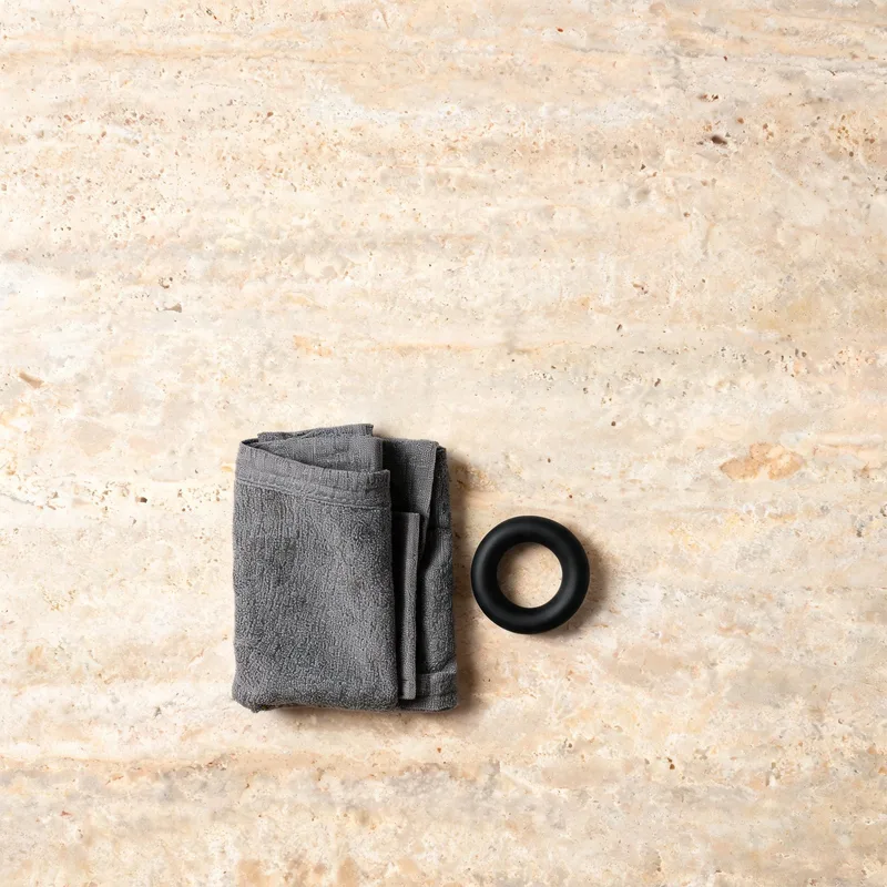 A discreet flatlay of a matte black silicone ring and a folded charcoal linen towel on a neutral stone surface, in soft diffused light