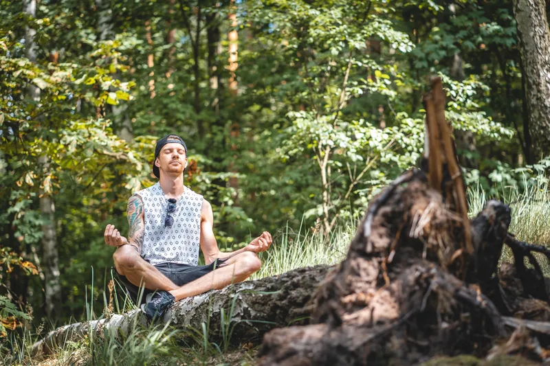 Man meditating peacefully with eyes closed while sitting on a fallen tree log in a sunlit forest