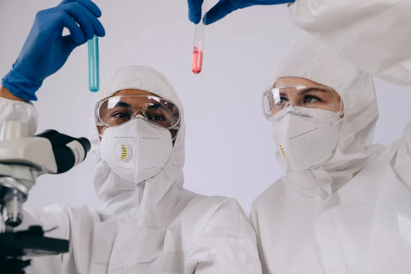 Two researchers in protective lab gear examining test tubes beside a microscope in a medical research laboratory